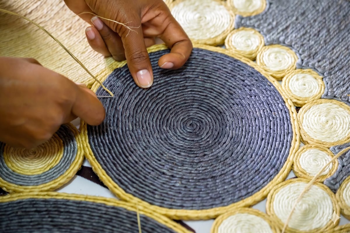 Black-and-white close-up of hands braiding pale sisal fibres, with the braid taking shape at the centre of the frame.